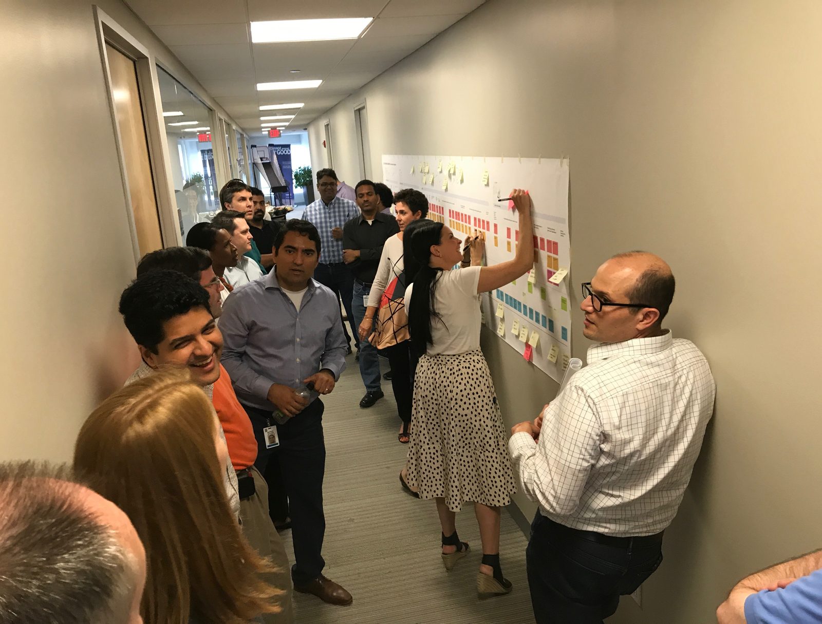 A crowd of colleagues gathered in a hallway around the service blueprint taped to the wall, one person reaching up to annotate it with a marker while others watch and point things out.