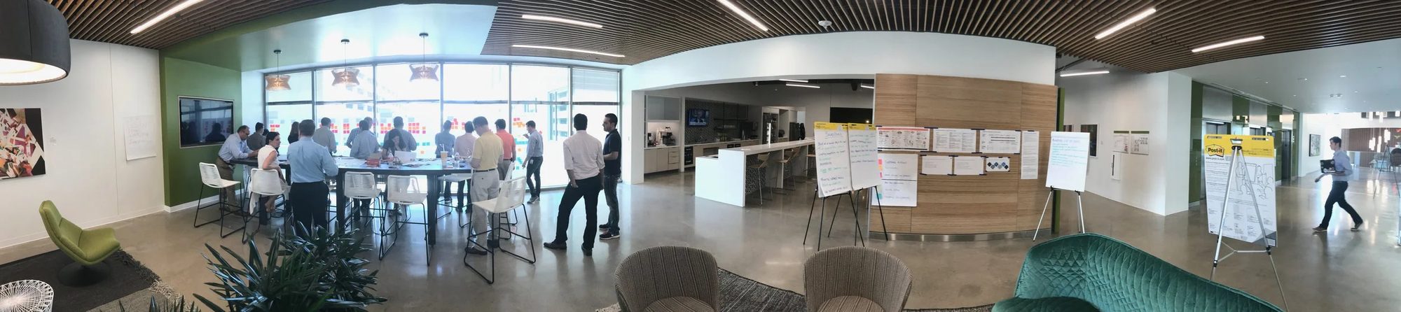 Wide panoramic view of an open office area near the kitchen, showing a team gathered around a table covered in sticky notes, with additional flip charts and working documents posted nearby.